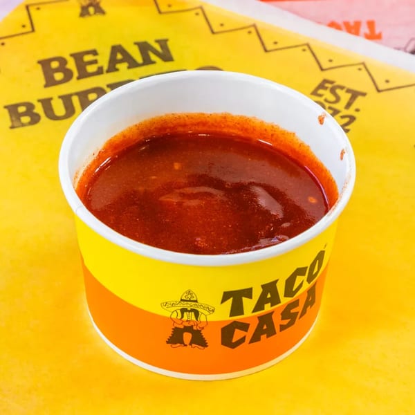 A Taco Casa branded cup filled with red chili sauce, on a yellow surface.