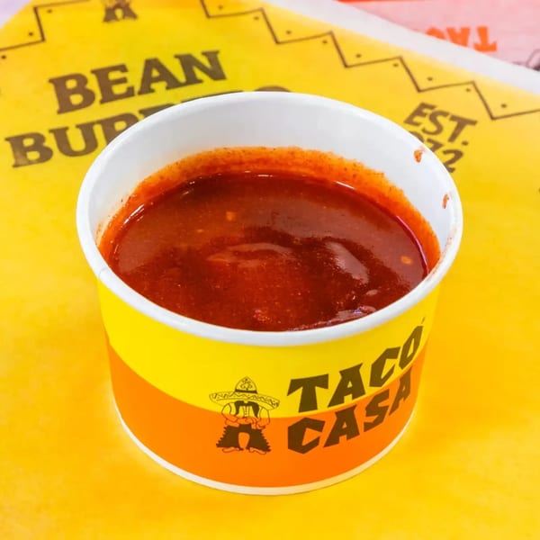 A Taco Casa cup filled with red chili sauce, on a yellow and pink background.