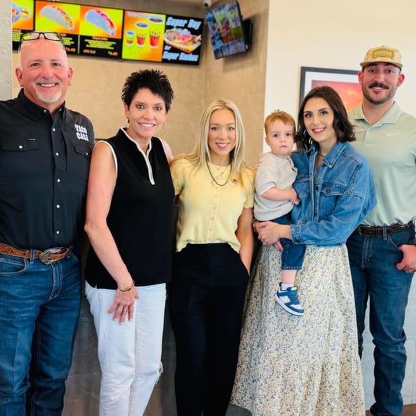 Three women, two men, and a baby smiling in front of a Taco Casa menu board.